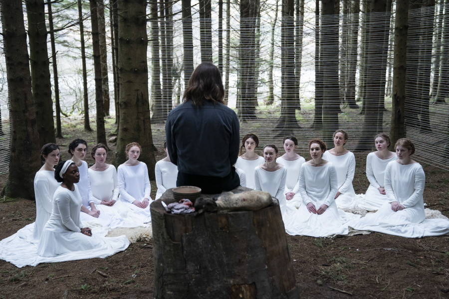 Shepherd (Michiel Huisman) sitting on a tree trunk in the forest. He is dressed in black and we see him from behind. In front of him on the ground sit about 15 women, all dressed in white, Selah (Rafe Cassidy) among them.