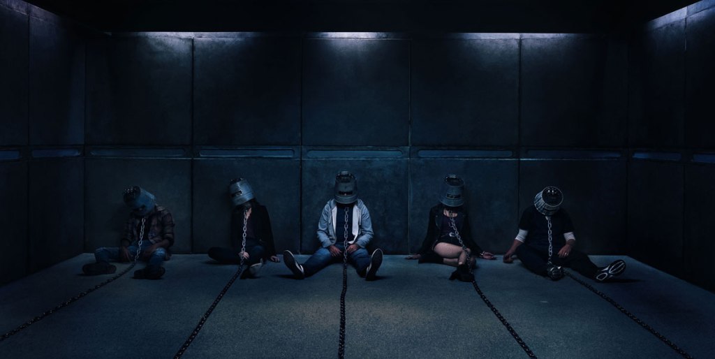 5 people with buckets over their heads and chains around their necks, slumped on the floor in an empty room.