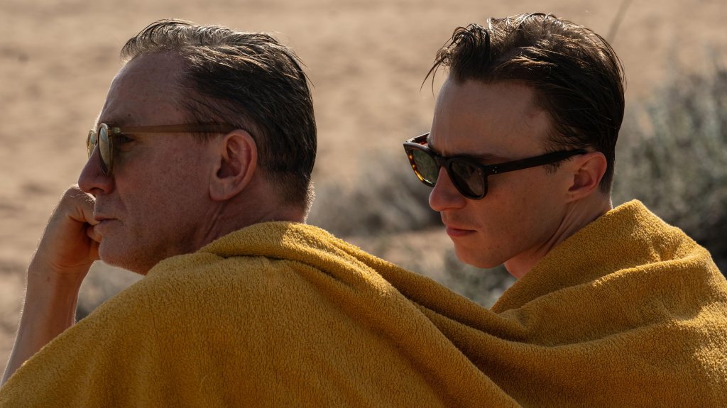 Lee (Daniel Craig) and Allerton (Drew Starkey) huddled under a towel on the beach. Lee is looking off into the distance, Allerton is leaning towards him. 