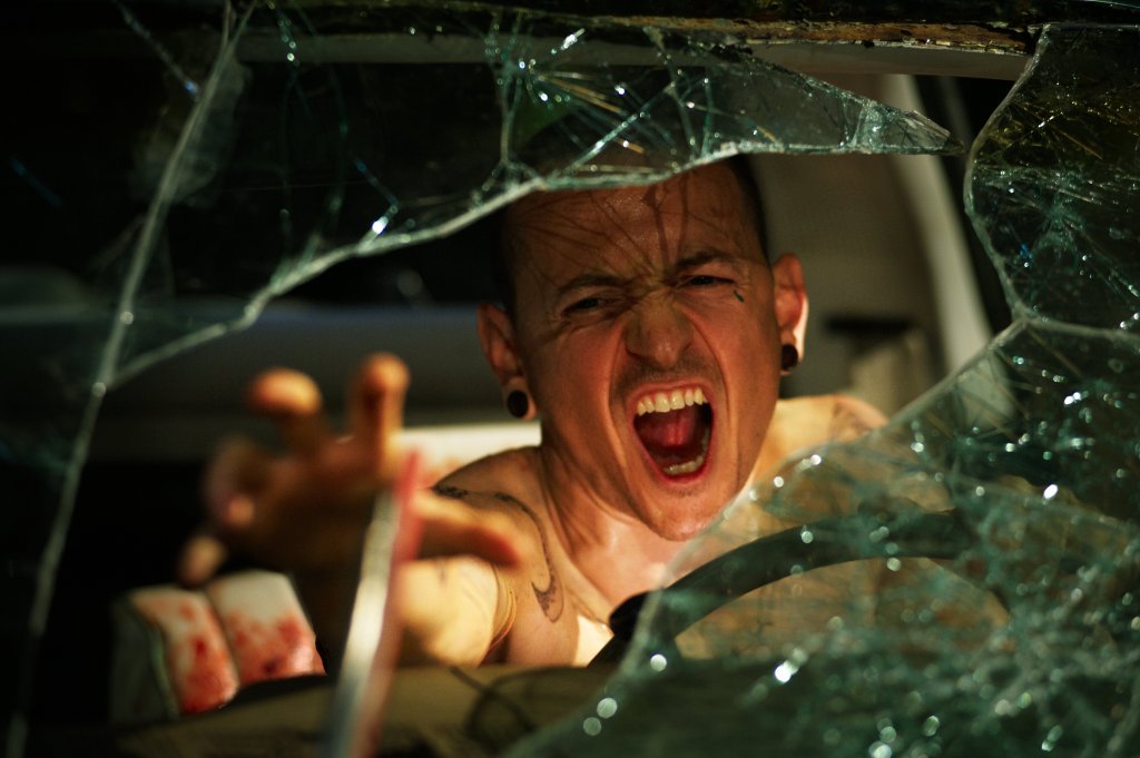 A nazi (Chester Bennington) in a car with a broken windshield reaching for something on the dashboard, screaming.