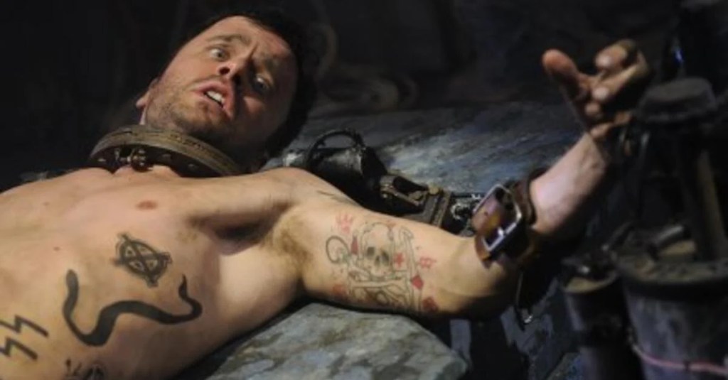 A shirtless man lying on a dirty table, his neck and hands are tied.