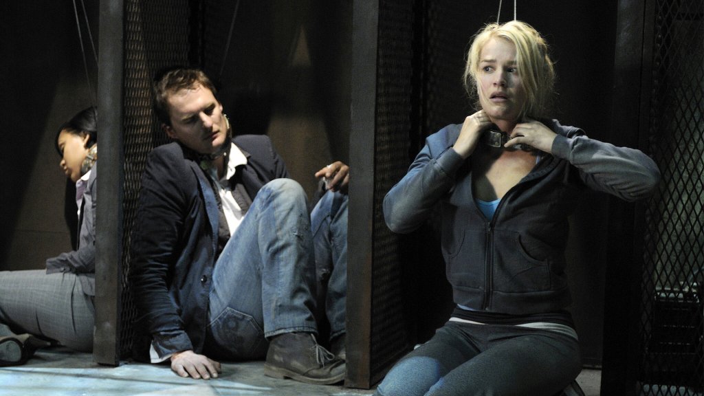 Three people huddled in open locker-like structures, wearing collars that are tied to the wall. 