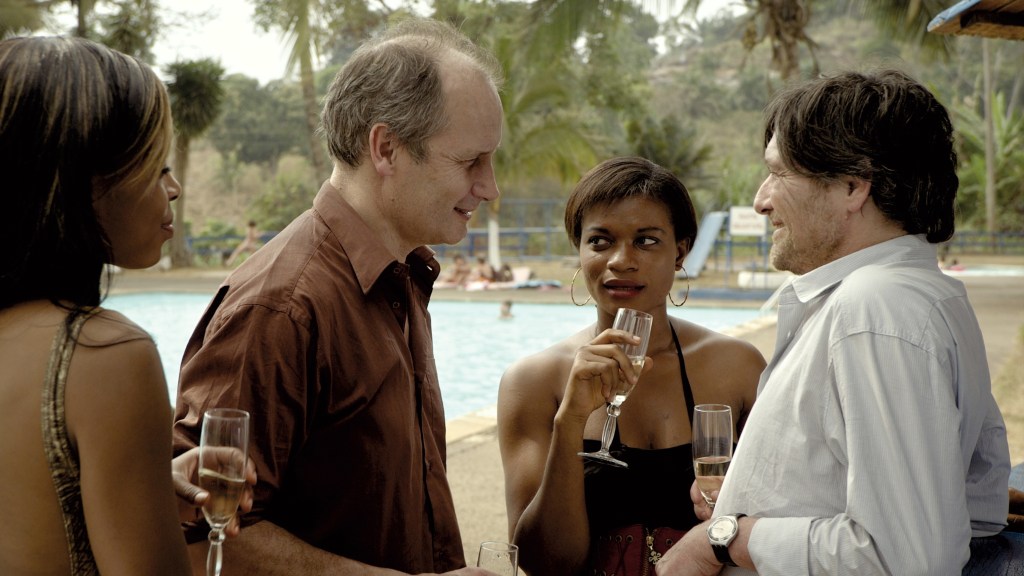 Ebbo (Pierre Bokma) leaning against a bar talking to another white guy. Two young Black women are standing next to them. Everybody is having champagne.