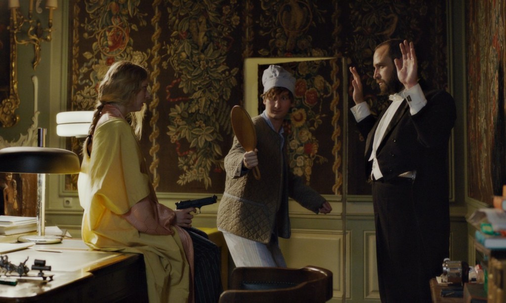 Octavia (Lilith Stangenberg) pointing a gun at Ljowushka (Aleksandre Koberidze) while Jakob (Alexander Herbst) holds up a mirror. He is dressed in his pyjamas.