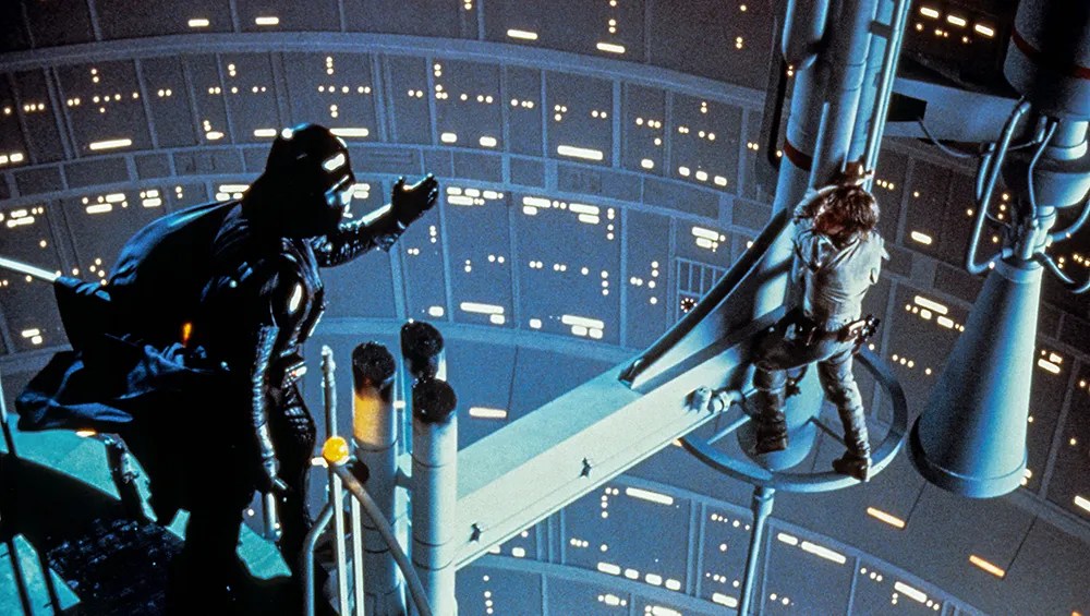 Darth Vader (David Prowse) reaching for Luke (Mark Hamill) who is just holding on to some machinery over emptiness.