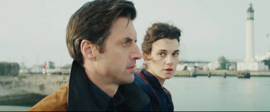 Oliver (Charlie Anson) and Agathe (Camille Rutherford) standing next to each other. He is looking in the distance, she is looking at him.