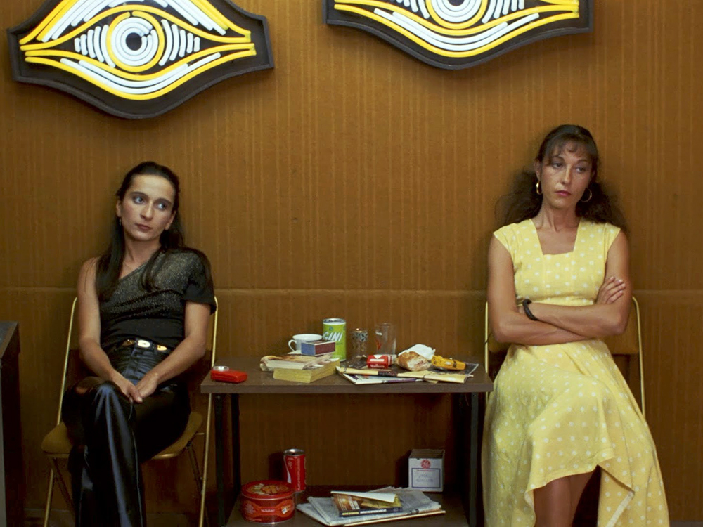 Simone (Ingrid Bourgoin) and Martine (Martine Simonet) sitting side by side with a table full of various trash between them, two glowing neon eyes on the wall above them.