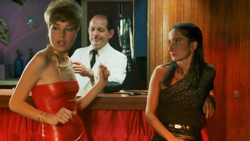 Simone (Ingrid Bourgoin) leaning against a bar, looking at a pretty young woman in a red dress. 