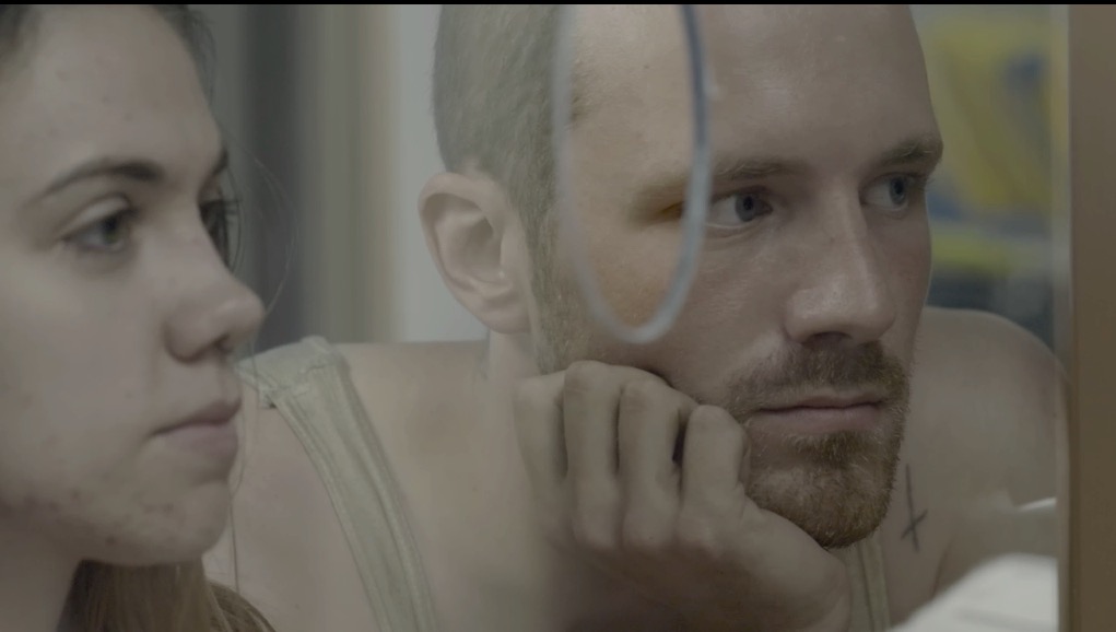 Blaise (Andrew Gillis) and Nessa (Bhreagh MacNeil) on opposite sides of a see-through plastic wall.