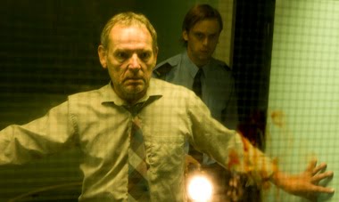 Robert Anderson (David Schofield) looking through a bloody window, a security guard behind him.