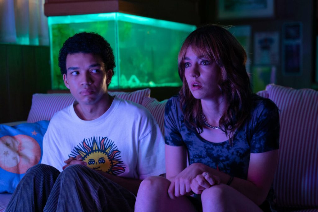 Owen (Justice Smith) and Maddie (Jack Haven) sitting on a sofa. Maddie is staring at the TV, Owen is looking slightly bewildered at her.