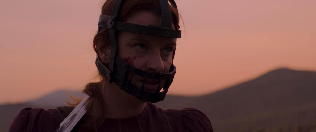 Ida Blye (Naomi McDougall Jones) in a metal mask that covers her mouth. 