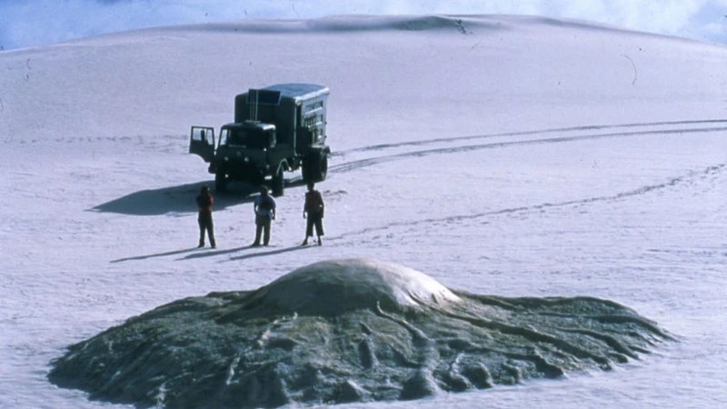 A truck stopped in the desert, three people standing before it, looking at some kind of alien organic structure.