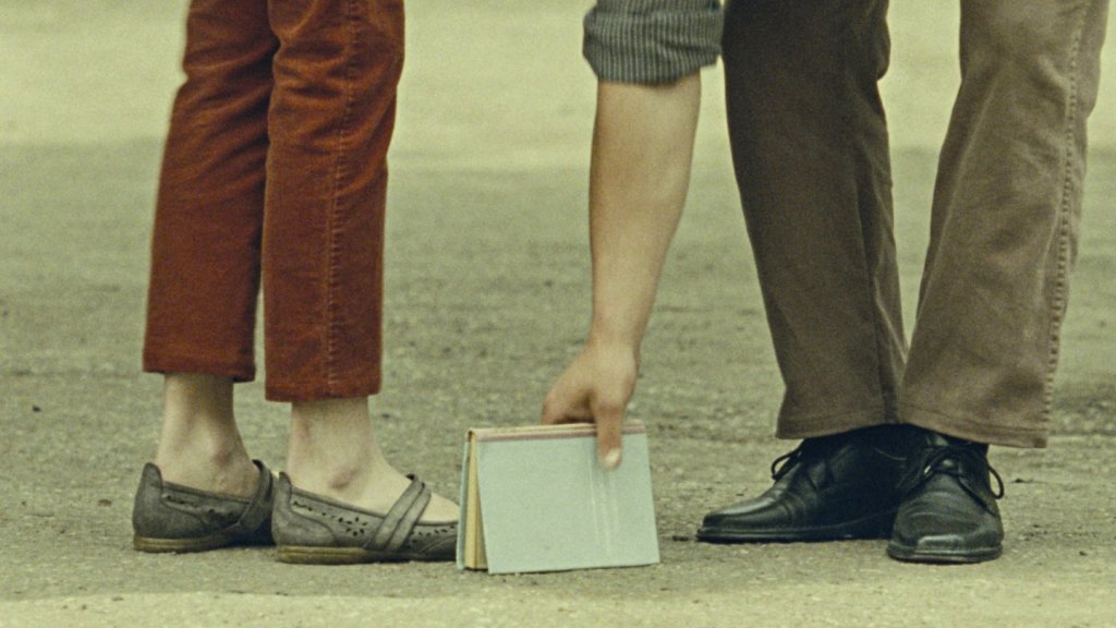 Two pairs of feet standing close to each other, as well as a hand grabbing a book that is lying on the floor.
