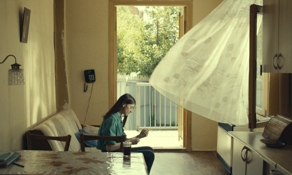 Lisa (Ani Karseladze) sitting in her apartment. The balcony door is open, as well as a window, the curtain is blowing into the room.