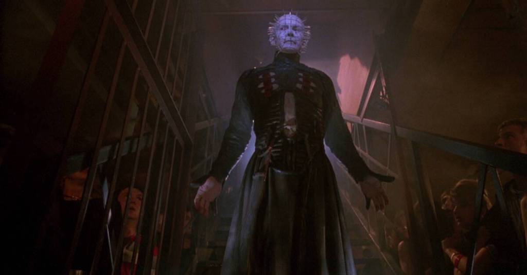 Pinhead (Doug Bradley) descending a staircase into a basement. Several people are hiding behind the stairs, watching him.
