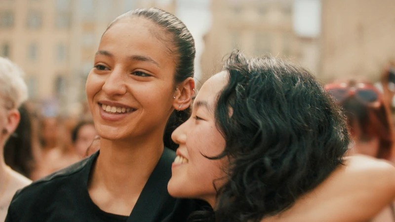 Fatima (Nadia Melliti) with her arm around Ji-Na (Park Ji-min) at a Pride parade, smiling broadly.