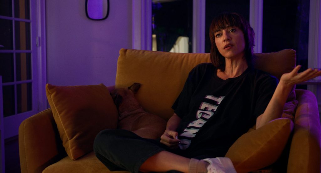 Ana Lily Amirpour sitting on a comfortable chair, a dog cuddled next to her, talking and gesticulating.