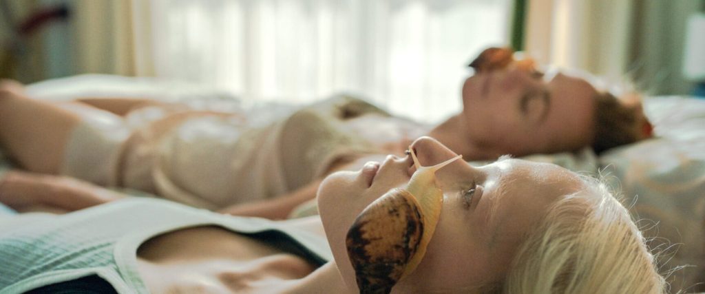 Masha (Marya Imbro) lying in bed next to her mother. Both have a white snail crawling over their faces.