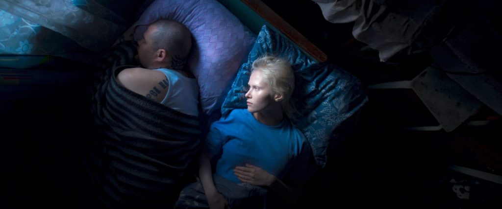 Masha (Marya Imbro) lying next to Misha (Mikhail Senkov). He is sleeping with his back turned to her, rolled up in a blanket. She is a awaka and looking at him.