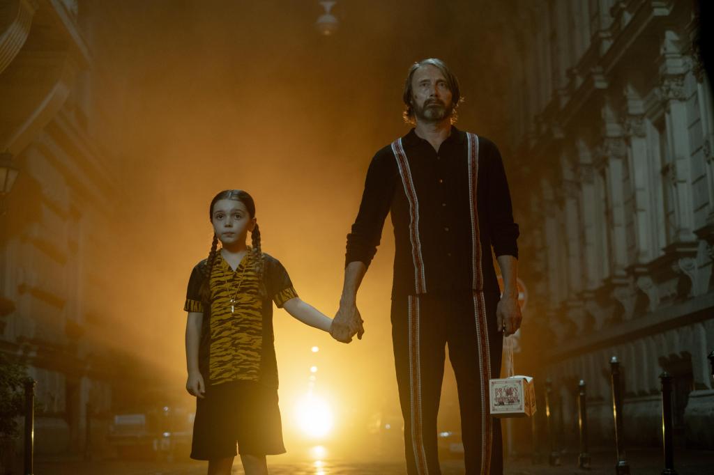 Aurora (Sophie Sloan) and the neighbor (Mads Mikkelsen) holding hands, standing in the middle of the street. He is holding take-out containers. 