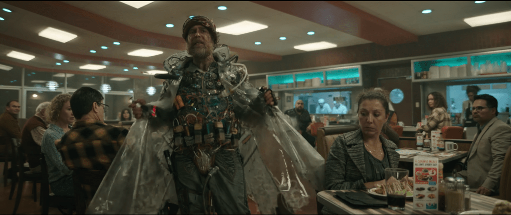A man (Sam Rockwell) in a diner. He is wearing some kind of technological vest that may or may not be a bomb and a plastic coat. 