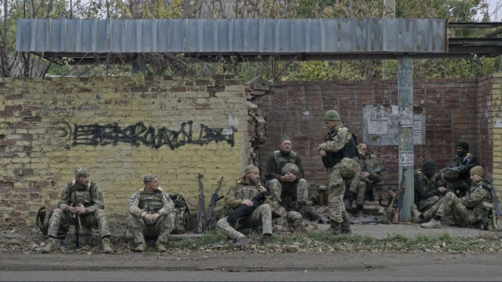 A group of soldiers at a ramshackle bus station, waiting. One of them has a dog on his lap. 
