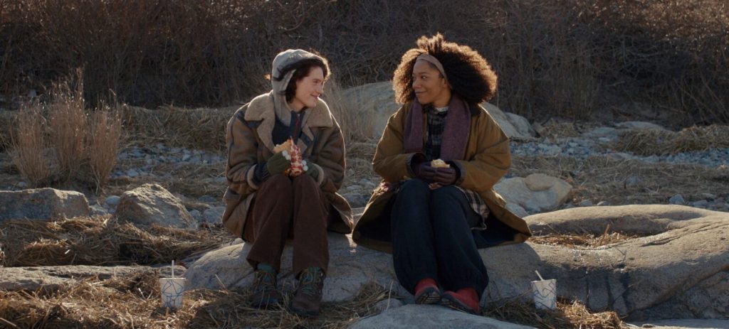 Agnes (Eva Victor) and Lydie (Naomie Ackie) sitting on a boulder, having lunch and smiling at each other. 