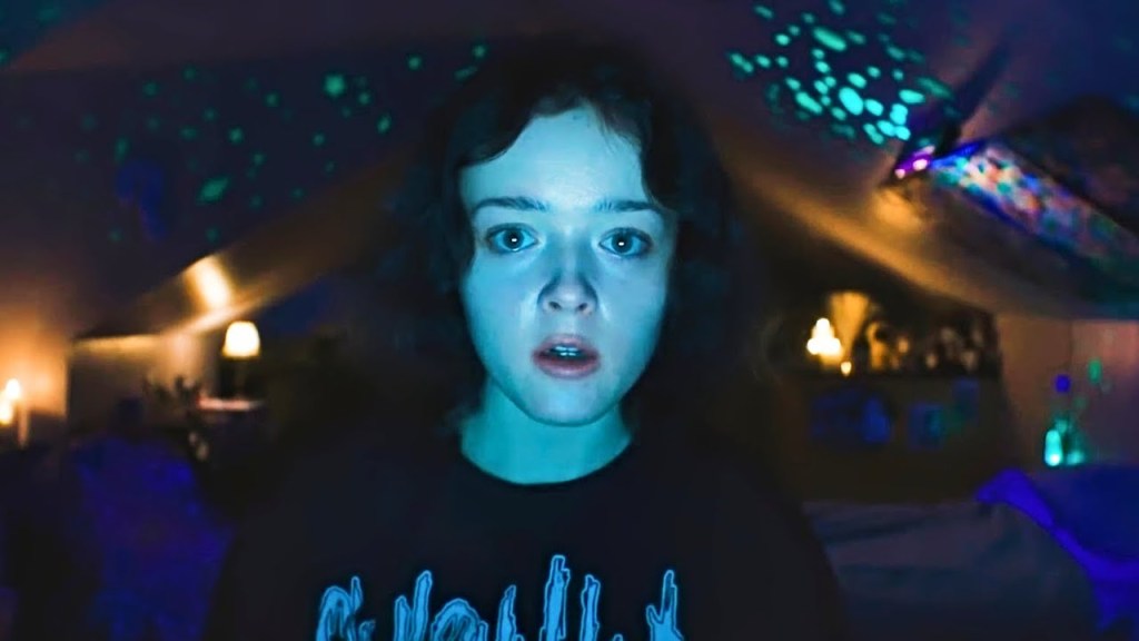Casey (Anna Cobb) looking at the camera of her computer screen. Her face is bathed in a blueish glow, behind her we can see glow in the dark stars on her slanted bedroom ceiling.