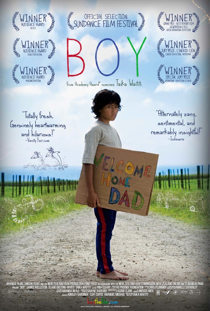 The movie poster showing Boy (James Rolleston) standing on a dirt road, holding a cardboard sign saying "Welcome Home Dad".