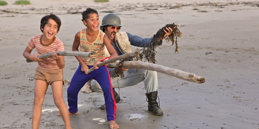 Rocky (Te Aho Eketone-Whitu), Boy (James Rolleston) and Alamein (Taika Waititi) playing on the beach. They are holding driftwood up as if it were big guns, screaming and laughing.