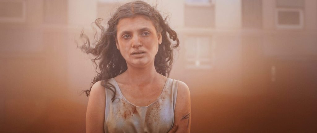 Alpha (Mélissa Boros) covered in dust, standing in what looks like a sandstorm with tears in her eyes.