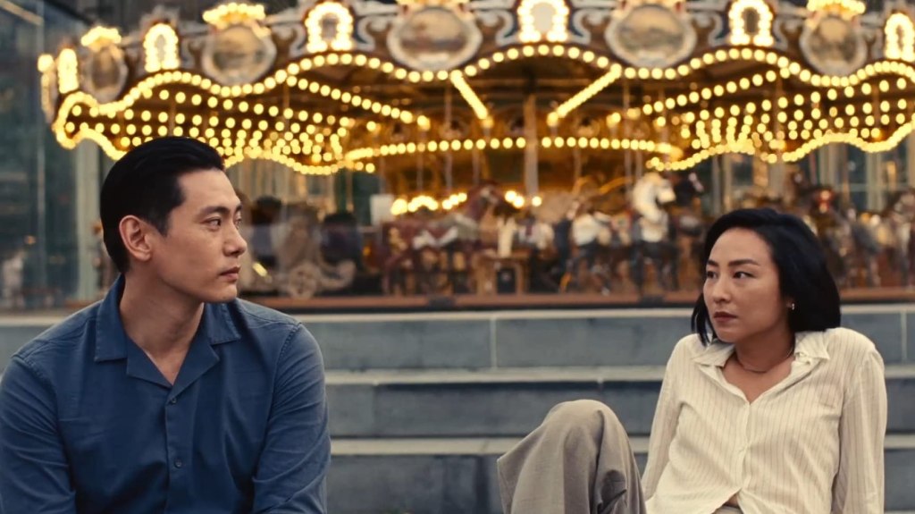 Hae Sung (Teo Yoo) and Nora (Greta Lee) sitting in front of an old merry-go-round, looking at each other.