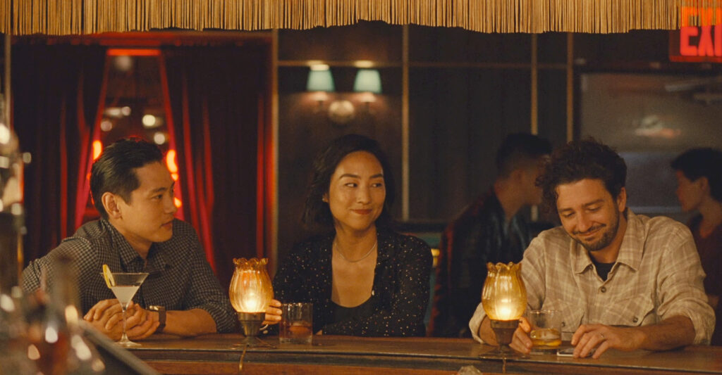Hae Sung (Teo Yoo), Nora (Greta Lee) and Arthur (Joe Magaro) sitting at a bar. Hae Sung and Nora are looking at Arthur, he is looking at his drink. Everybody is smiling.
