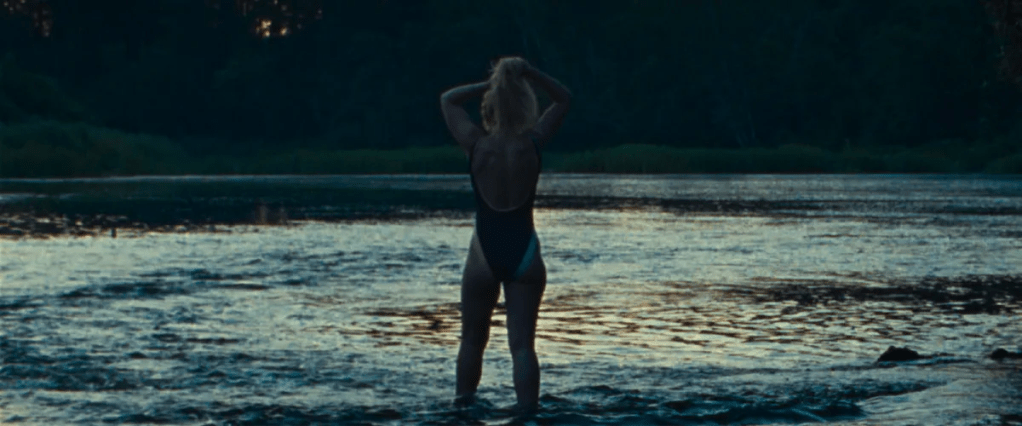 Lidia (Imogen Poots) standing in a lake that reaches to her ankles. The sun is setting, it is almost dark. She is holding her hair up.