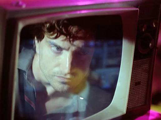 An old TV showing a man looking very intently, overlayed by what appears to be a news program. It is unclear what is inside the TV and what is the reflection. 