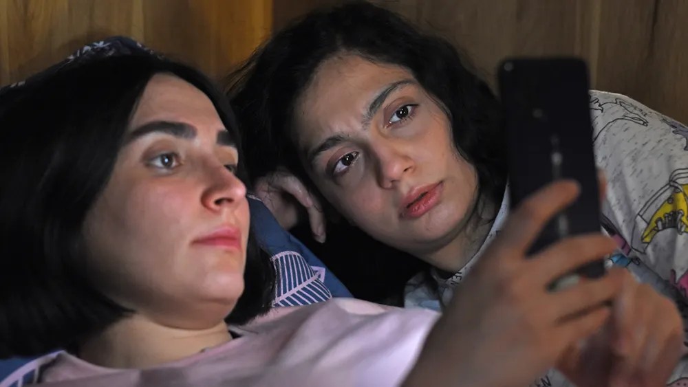 Rezvan (Mahsa Rostami) and Sana (Setareh Maleki) lying in bed together and looking at a smartphone screen with very serious expressions.