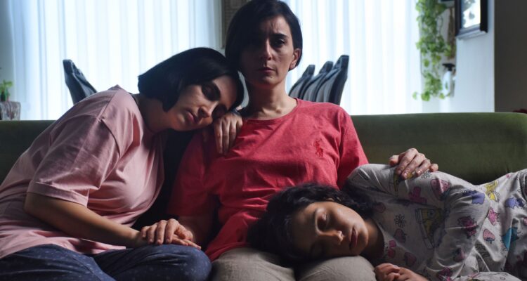 Najmeh (Soheila Golestani) cuddling Rezvan (Mahsa Rostami) and Sana (Setareh Maleki) on the couch. The two daughters are sleeping, while Najmeh looks ahead with a serious expression.