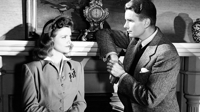 Irena (Simone Simon) and Oliver (Kent Smith) leaning against the mantle of a fireplace, looking at each other.