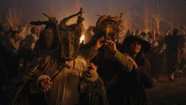 Two people with rustic wood masks. Behind them is a large group of people around a bonfire.