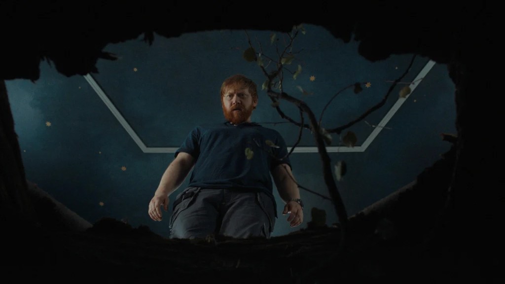 Jon (Rupert Grint) looking down a hole in the floor of the kid's room. A small tree is growing from it. 