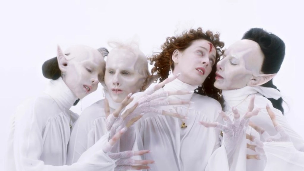 The four vampire sisters in a music video. They are all dressed in white, and the background is also white. Three are in vampiric forms with bald heads and elven ears, one in human form. All have elongated fingers though.