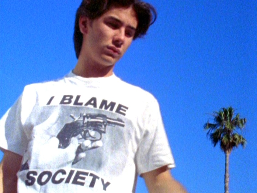 Andy (James Duval) wearing a t-shirt with hands that are holding up a small revolver. It says "I blame society" in big black letters.