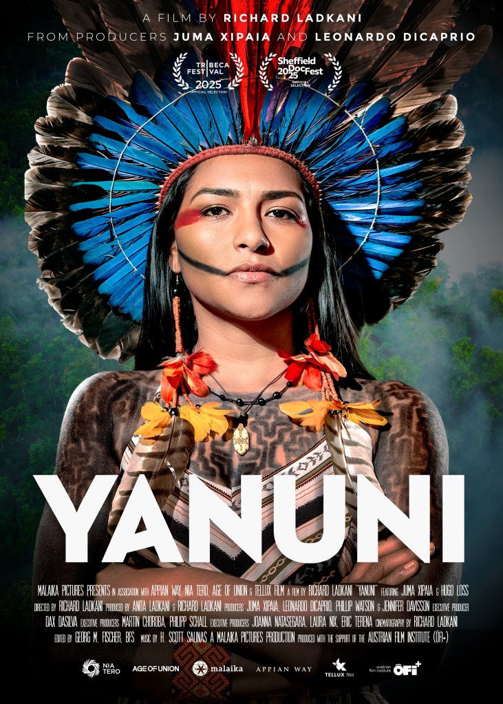 The movie poster showing Juma Xipaia in a traditional headdress made of feathers. She has equally traditional make-up and feather earrings, markings on her chest and upper arms that might be tattoos and a dress. She is looking straight at the camera, her arms crossed over her chest.