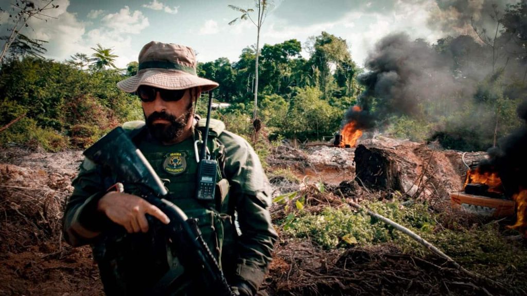 Xipaia's husband walking away from burning mining equipment in the Amazon. He has a rifle in his hands and is wearing military garb.