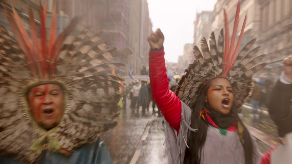 Juma Xipaia at a protest, waring a differnt traditional headdress, her fist raised. She seems to be shouting something.