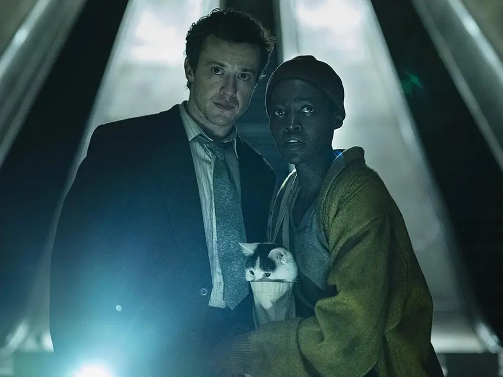 Eric (Joseph Quinn) and Sam (Lupita Nyong'o) at the bottom of some escalators. It is dark except for a torch that Eric is holding. Sam has her cat in her bag.
