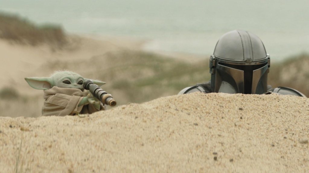 Grogu and Mando hiding peeking over a sanddune. Grogu is looking through a looking glass.