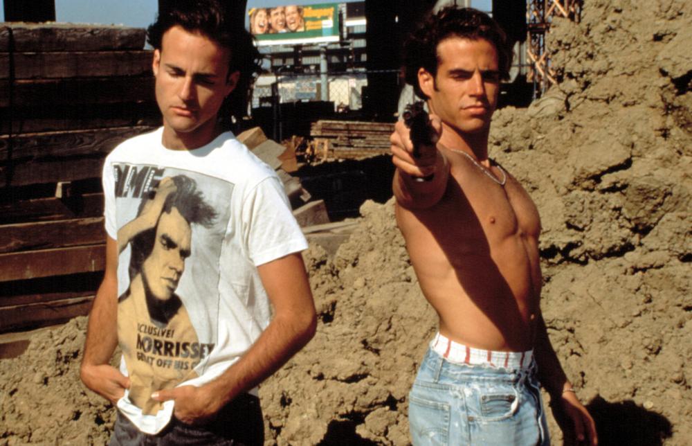 Jon (Craig Gilmore) and Luke (Mike Dytri)s standing almost back to back. Jon has his eyes closed. Luke is shirtless and pointing a gun towards the camera.