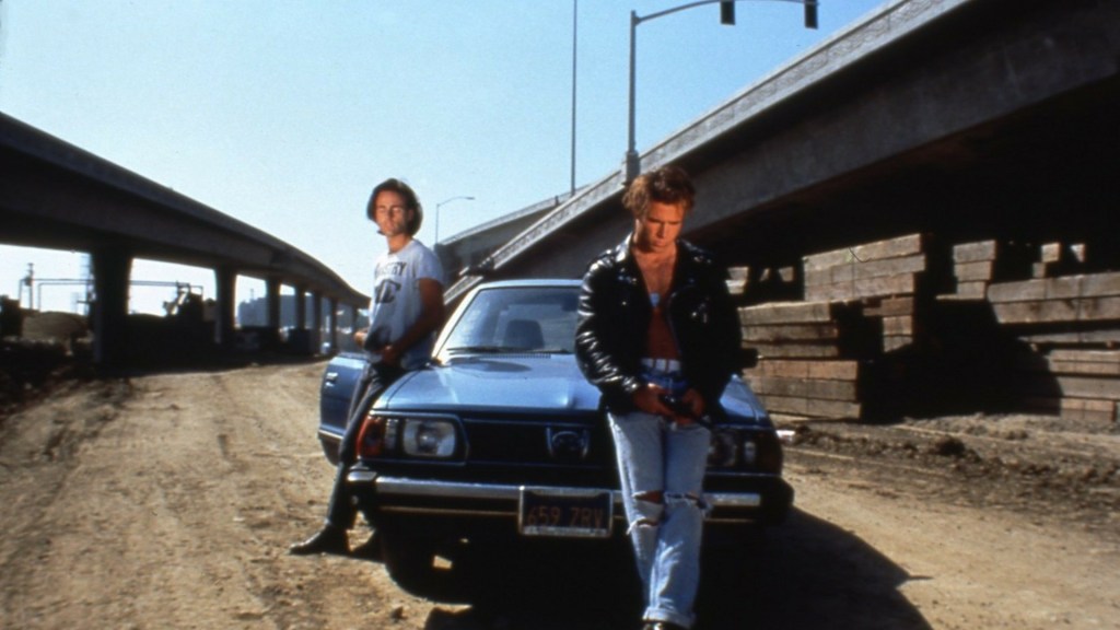 Jon (Craig Gilmore) and Luke (Mike Dytri) leaning against a car parked underneath an overpass, next to construction material.